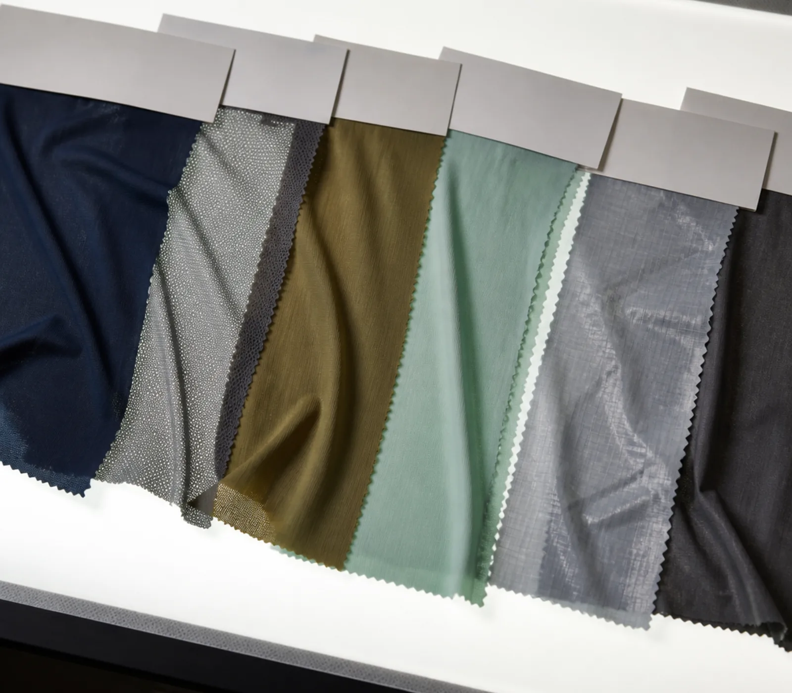 Seven fabric swatches of different weights and finishes hanging from a plain grey swatch header