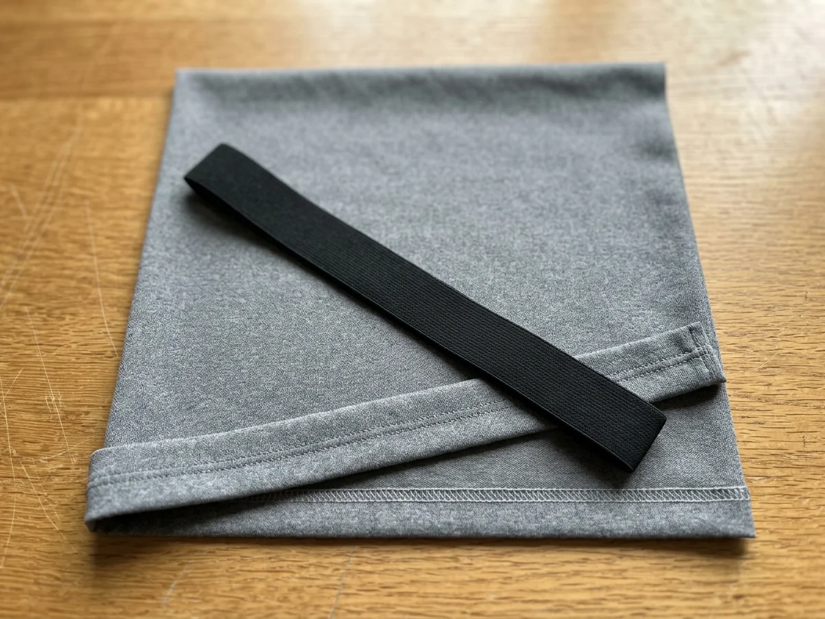 A panel of grey performance knit with a coverstitched edge lying on an oak desk, a length of flat elastic band laid across it